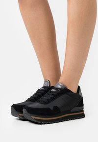 Baskets noires en suede et matière synthétique, accents gris texturés, fermeture à lacets et semelle en caoutchouc avec un motif rayé.