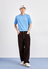 Camiseta de algodón azul claro con un logo, pantalones de pana marrones y zapatillas blancas con detalles en beige, sobre un fondo liso.