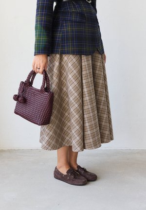 Persoană purtând o fustă și o jachetă în carouri, ținând o geantă violetă țesută și purtând pantofi maro cu șireturi, stând pe un podea deschisă la culoare.
