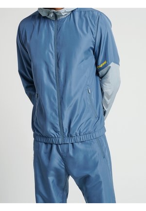 Mand iført blå træningsjakke med lynlås og matchende bukser med lysegrå ærmer og gul branding på armen.