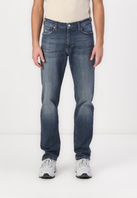 Jean en denim bleu avec une jambe droite, un design classique à cinq poches, une texture délavée et un petit patch logo sur la poche avant.