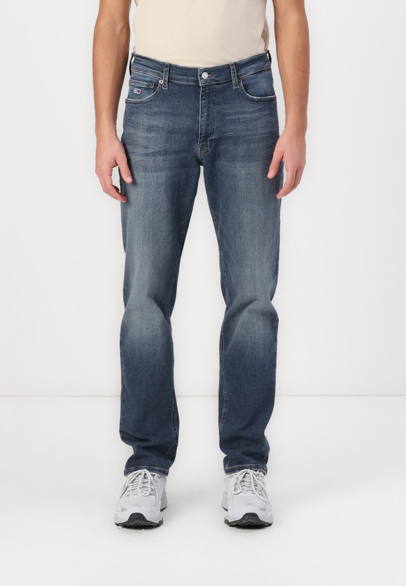 Jean en denim bleu avec une jambe droite, un design classique à cinq poches, une texture délavée et un petit patch logo sur la poche avant.
