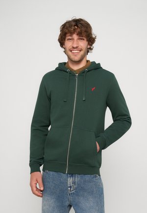 Junger Mann lächelt, trägt einen dunkelgrünen Zip-Hoodie mit Vordertaschen und blaue Jeans, steht vor einem schlichten hellen Hintergrund.