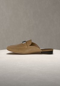 Mocassin marron en cuir à enfiler avec un détail de boucle argentée, talon plat et bout arrondi, reposant sur une surface claire sur un fond neutre.
