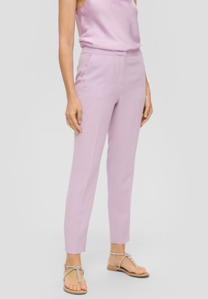 Femme portant un pantalon fuselé violet clair, un top sans manches assorti et des sandales plates beige avec des sangles à la cheville, debout devant un fond uni.