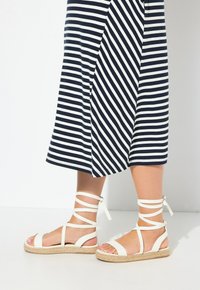 Białe sandały na cienkich paskach z espadrylową podeszwą i wstążkowymi akcentami, w połączeniu z granatową sukienką w biało-niebieskie paski.