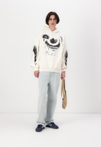 Kremhette med svart abstrakt design, kombinert med lyseblå jeans og marineblå sko. Holder en brun veske over den ene skulderen.