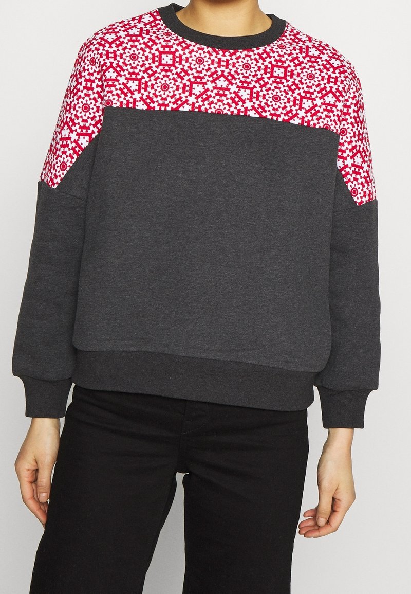 Personne portant un sweat-shirt gris foncé avec un motif géométrique rouge et blanc sur les épaules et le haut de la poitrine, associé à un pantalon noir.