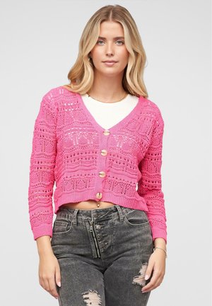 Cardigan rose en tricot avec un col en V, fermeture à boutons sur le devant et un design ajouré complexe. Associé à un jean foncé déchiré.