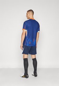 Blå sports trøje med orange og lyseblå accenter, parret med marineblå shorts. Sorte knæhøje sokker og hvide og sorte fodboldstøvler.