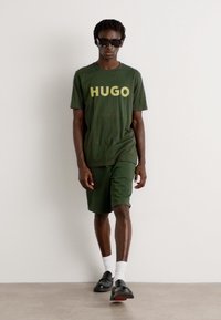 Grøn bomulds t-shirt med "HUGO" i gul tekst, parret med matchende grønne shorts. Model med solbriller og sorte sko med hvide sokker.