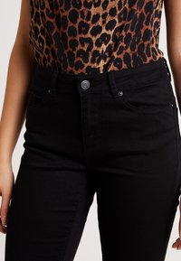 Kvinna som bär högmidjade svarta jeans och en ärmlös leopardmönstrad topp mot en enfärgad bakgrund.