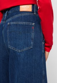 Person som har på seg mørkeblå Replay-jeans med brun lærmerke og rød langermet skjorte, som viser baklomme og sømdetaljer.