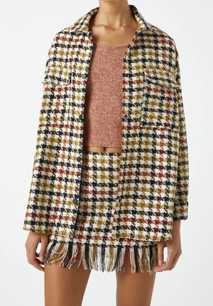 Kvinde iført en tekstureret ternet jakke med knapper og et matchende frynset skørt, kombineret med en cropped støvet lyserød strikket top.