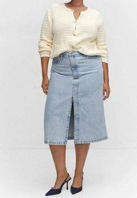 Personne portant un cardigan en maille crème avec des boutons dorés, une jupe midi en denim bleu clair avec une fente à l'avant, et des talons pointus bleu marine.