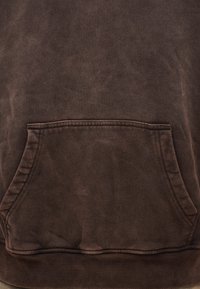 Κοντινή λήψη μπροστινής τσέπης τύπου καγκουρό σε καφέ φούτερ με ορατές ραφές και απαλό, φθαρμένο υφασμάτινο υφή.