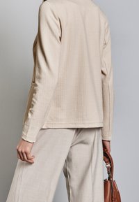 Béžová texturovaná bunda s dlouhými rukávy, pohled zezadu. Kombinována s kalhotami světlé barvy a hnědou kabelkou, zdůrazňující jednoduchý, čistý design.