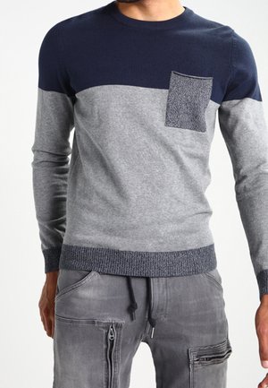Mann trägt einen farbblockierten Pullover mit dunkelblauem Oberteil, grauem Mittelteil, texturierter Brusttasche und grauen Cargohosen mit Reißverschlusstaschen.