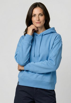 Frau mit schulterlangem braunem Haar, die einen hellblauen Kapuzenpullover und dunkle Hosen trägt, posiert mit einer Hand nahe ihrem Kinn vor einem schlichten Hintergrund.