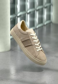 Μπεζ αθλητικό παπούτσι από σουέτ με λευκά κορδόνια και σόλα, που αιωρείται πάνω από ένα γυαλισμένο δάπεδο από σκυρόδεμα κοντά σε έναν τοίχο από γυάλινα τούβλα.