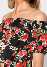 Femme portant un haut noir à épaules dénudées avec un motif floral rouge, blanc et orange et des manches courtes bouffantes.