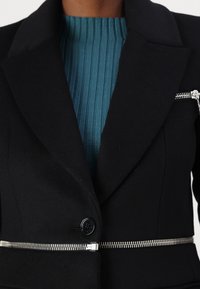 Blazer in lana nera con colletto appuntito, chiusura a bottoni e dettaglio in zip argentato lungo la vita. Sotto, un maglione a coste color teal.