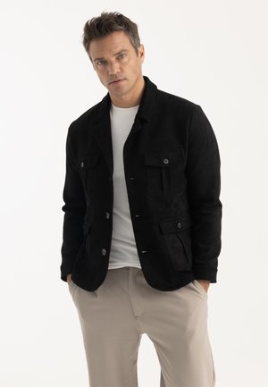 Homme aux cheveux courts portant une veste noire boutonnée sur une chemise blanche et un pantalon beige, les mains dans les poches, sur un fond uni.