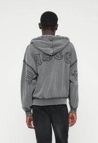 Hanorac gri cu finisaj uzat, croială oversized și broderie cu litere pe spate. Dispune de manșete elastice și talie elastică, textură netedă.