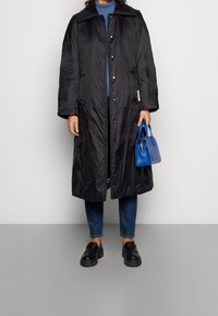 Černý dlouhý prošívaný kabát s límcem, patentkami a bočními kapsami. Spárován s modrým topem a džíny. Drží malou modrou kabelku.