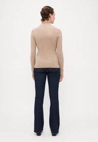 Beige long-sleeve top with a high neckline, fitted design, and ribbed hem, paired with dark blue flared jeans and black footwear.