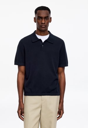 Jeune homme portant un polo noir à manches courtes par-dessus un t-shirt blanc à col rond, associé à un pantalon beige, debout devant un fond blanc.