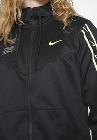 Černá bunda na zip s kapucí, s neonově zelenými akcenty a logy na hrudi a rameni. Vyrobená z hladkého, lehkého materiálu.