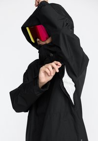 Svart vattentät jacka med dragsko i huvan, dragkedjeventilation och texturerad foder. Modellen bär reflexglasögon.