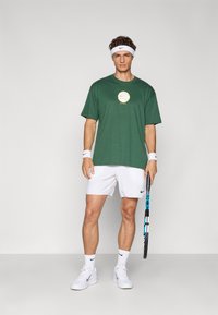Grøn kortærmet Nike t-shirt med rundt logo, hvide tennis shorts, hvide sneakers og håndledsbeskyttere; holder en tennisracket med blå accenter.