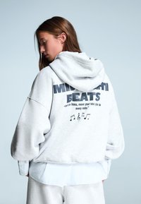 Sivý prehnaný mikina s kapucňou, so čiernym nápisom "MIDNIGHT BEATS" a hudobnými notami na zadnej strane, vyrobená z mäkkej zmesi bavlny.