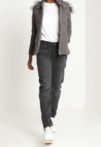 Grå dunjakke med pelsforet hætte, hvid skjorte under, stramme sorte jeans og hvide sneakers. Gående stilling mod en lys baggrund.