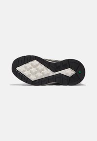 Suela de zapatilla en blanco y negro con agarre texturizado, patrones de huella en forma de cruz y un pequeño hexágono verde cerca del talón sobre un fondo blanco.