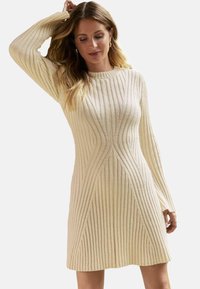 Robe pull tricotée crème avec un motif à côtes verticales, manches longues et un corsage ajusté avec un ourlet évasé.