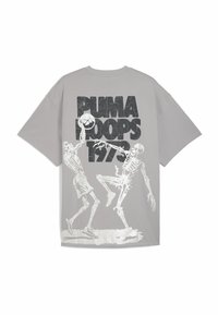 Camiseta gris de gran tamaño con un gráfico de esqueleto blanco jugando al baloncesto, con texto en negrita negro "PUMA HOOPS 1973" arriba. Material de algodón.