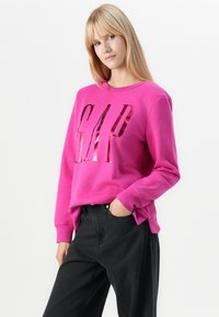 Fuksia sweatshirt med en blank "GAP" logo i fet skrift. Lange ermer med ribbede mansjetter, kombinert med mørk svart denim bukser.
