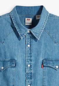 Hele sinine teksasärk, millel on terav krae, eesmine lett hõbedaste nööpidega, kaks rinnataskut ja väike punane Levi'si silt.