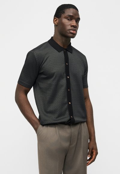 Νέος άνδρας με κοντά μαλλιά, φορεμένος με μαύρο πουκάμισο με σχέδιο και κοντά μανίκια και μπεζ παντελόνια, στέκεται με το ένα χέρι στην τσέπη.