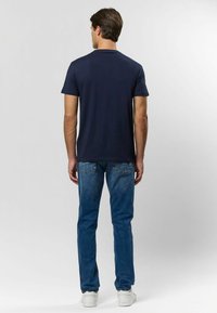 Námořnická modrá tričko s krátkým rukávem a kulatým výstřihem, nosené s světle modrými džíny a bílými teniskami, viděné z bat.