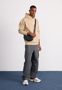 Felpa beige con tasca a marsupio, pantaloni cargo grigi e scarpe sportive bianche con dettagli viola. Borsa a tracolla nera con cintura regolabile.
