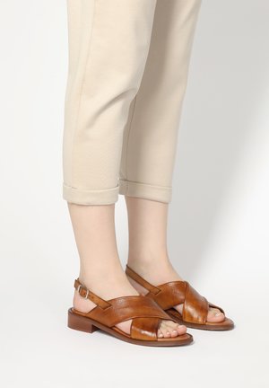 Brown leather sandals with cross-strap design, open toe, and ankle strap. Features a low block heel and smooth texture.
