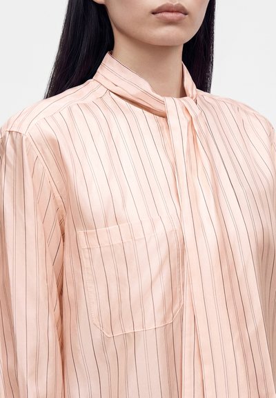 Femme portant une blouse rose clair à fines rayures avec un col montant et une poche avant, vue du cou au milieu du torse.