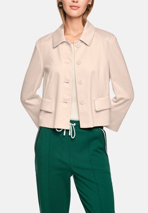 Woman wearing a beige buttoned jacket with flap pockets, white top, and green drawstring pants with white side stripes, hands behind back.