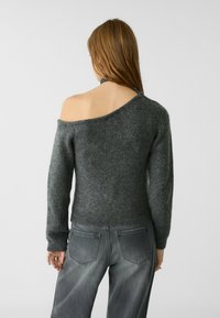 Asymmetrischer, dunkelgrauer Pullover mit strukturiertem Strick, mit einem schulterfreien Design und langen Ärmeln, kombiniert mit hellgrauen Jeans.