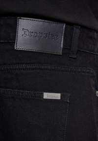 Gros plan de la poche arrière d’un jean en denim noir avec une étiquette en cuir cousue et une plaque métallique, toutes deux embossées avec le mot "Dropsie."