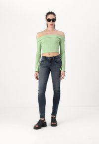 Vállra tett, bordázott zöld crop top; sötét skinny farmernadrág; fekete blokk-sarkú szandál; minimális kiegészítők; sima szövettextúra.